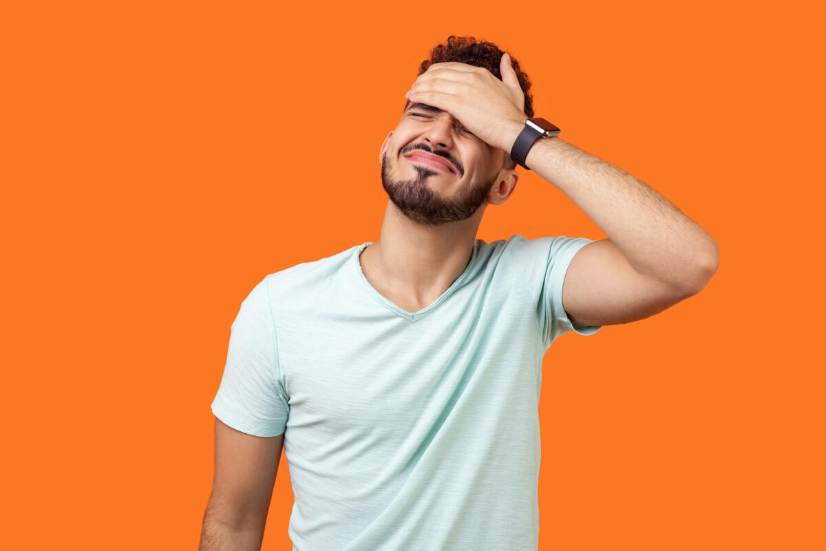 Porträt eines verzweifelten brünetten Mannes mit Bart in weißem T-Shirt, der das Gesicht mit der Hand bedeckt. © Khosrork / Getty Images