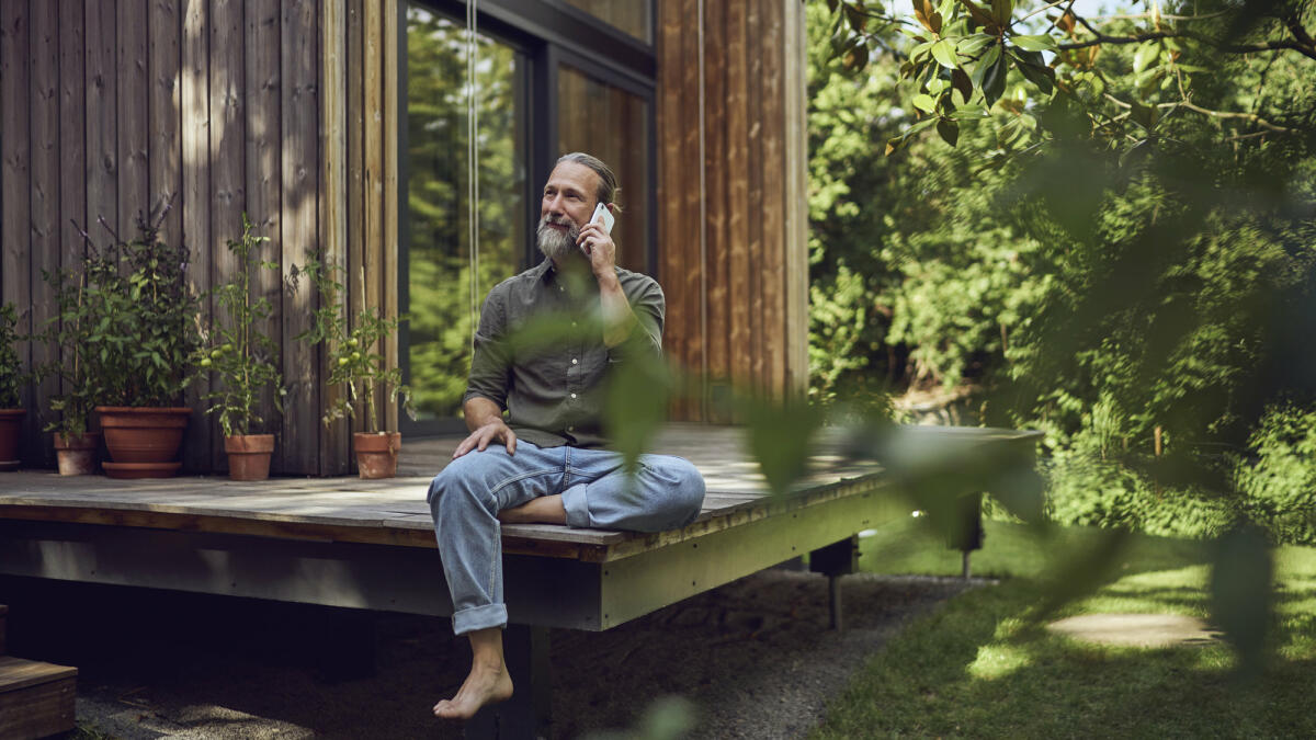 Ein älterer Mann mit grauen Haaren und Zopf sitzt auf einer Terrasse im Grünen und telefoniert lächelnd. © Westend61 / Getty Images