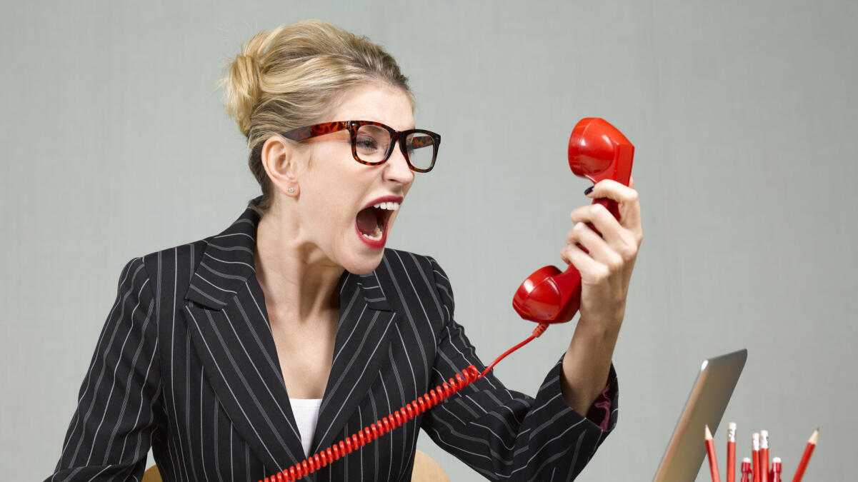 Wütenden Businessfrau sitzt an ihrem Schreibtisch und schreit in das Telefon © Peter Dazeley / Getty Images