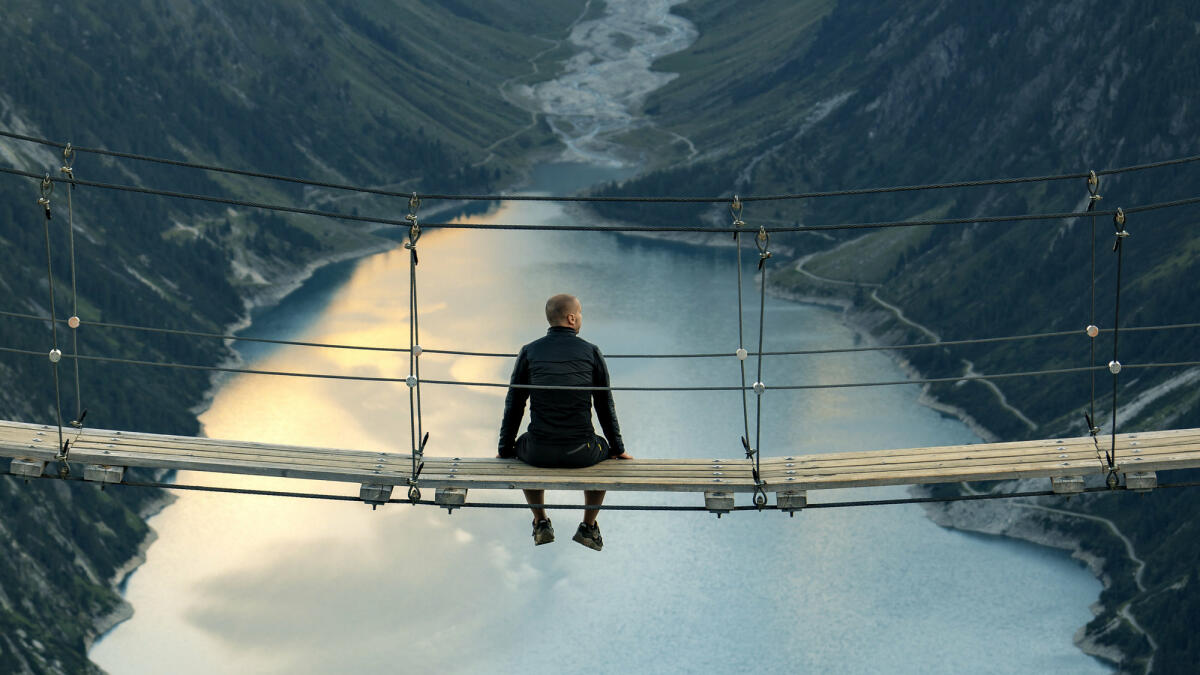 Rückansicht des Mannes, der auf einer Seilbrücke sitzt und die Berglandschaft betrachtet. © ljubaphoto / Getty Images