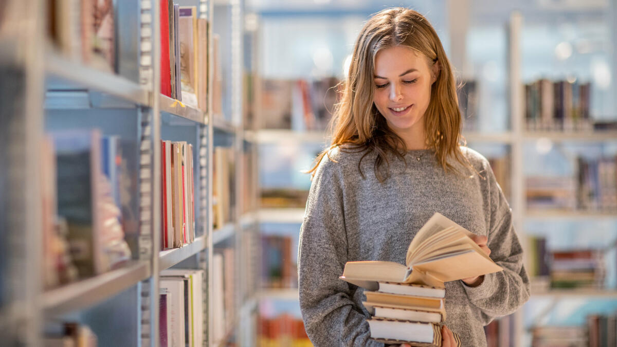 Frau steht in der Bibliothek mit einem Buch in der Hand vor dem Regal © Lorado / Getty Images