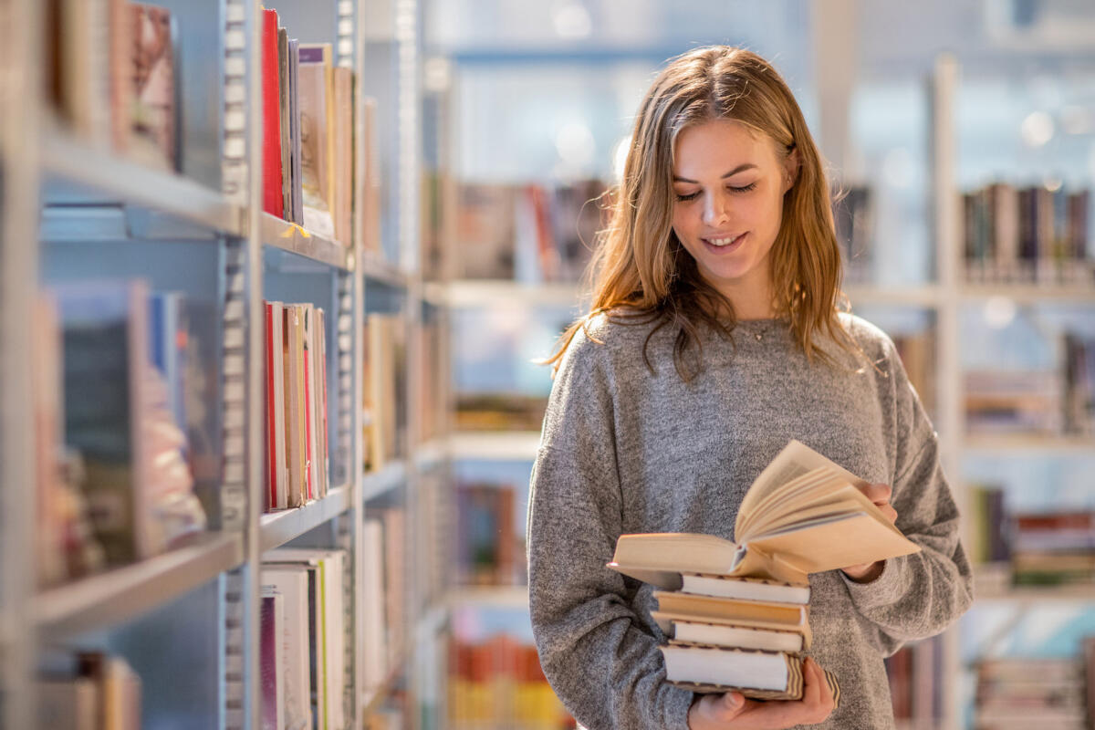 Frau steht in der Bibliothek mit einem Buch in der Hand vor dem Regal &copy; Lorado / Getty Images