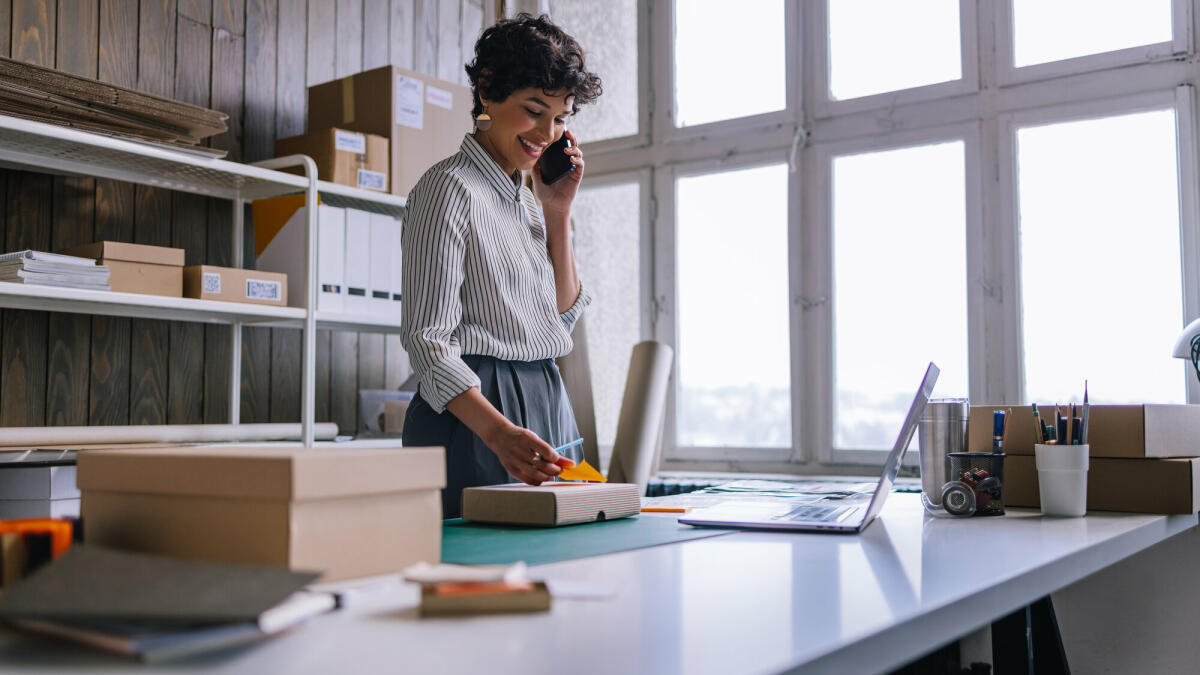 Eine schöne, lächelnde lateinamerikanische Unternehmerin, die ihr Handy benutzt, während sie Waren für die Lieferung in Kartons verpackt. © Getty Images/ FreshSplash