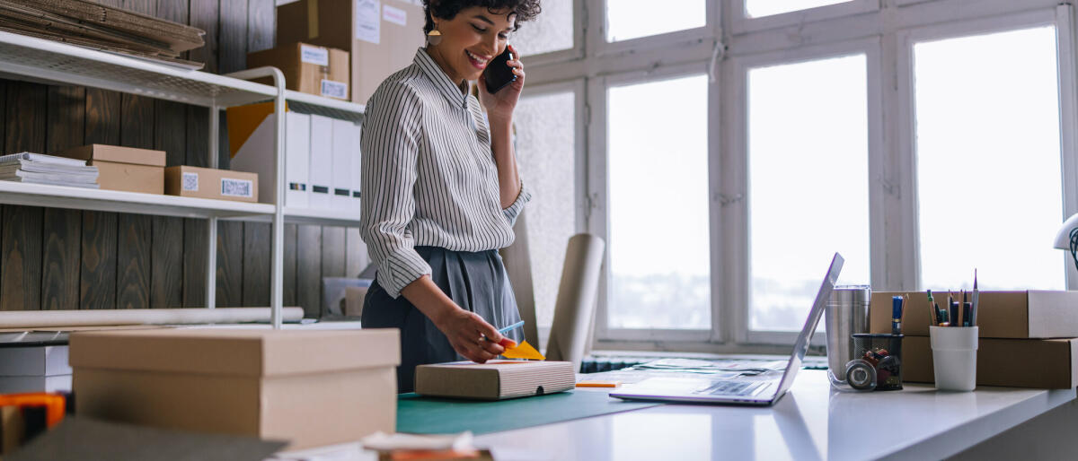 Eine schöne, lächelnde lateinamerikanische Unternehmerin, die ihr Handy benutzt, während sie Waren für die Lieferung in Kartons verpackt. © Getty Images/ FreshSplash