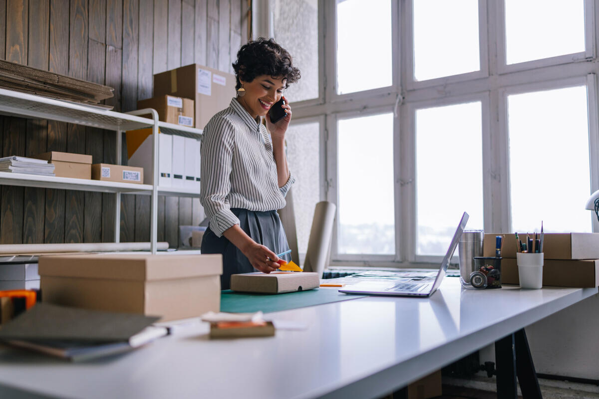 Eine schöne, lächelnde lateinamerikanische Unternehmerin, die ihr Handy benutzt, während sie Waren für die Lieferung in Kartons verpackt. © Getty Images/ FreshSplash