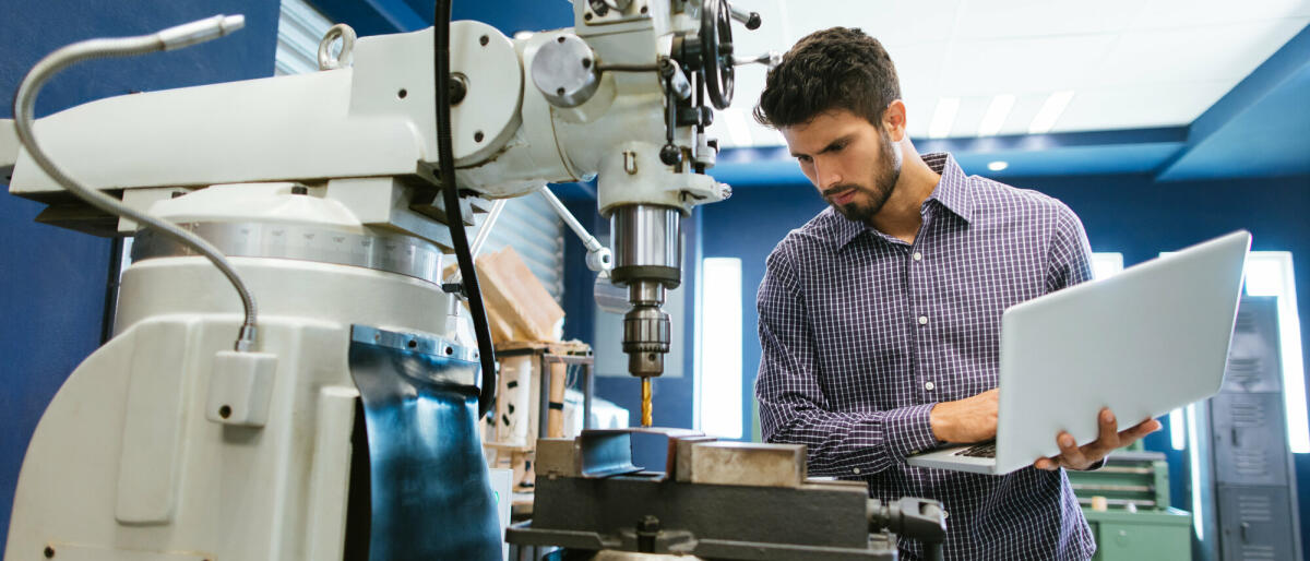 Ein junger lateinamerikanischer Ingenieur kalibriert eine große Bohrmaschine mit seinem Laptop in seiner Werkstatt. © aldomurillo/ Getty Images 