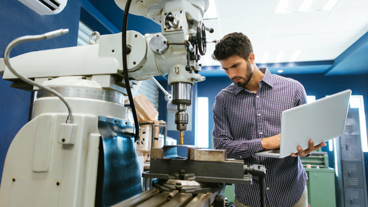 Ein junger lateinamerikanischer Ingenieur kalibriert eine große Bohrmaschine mit seinem Laptop in seiner Werkstatt. © aldomurillo/ Getty Images 