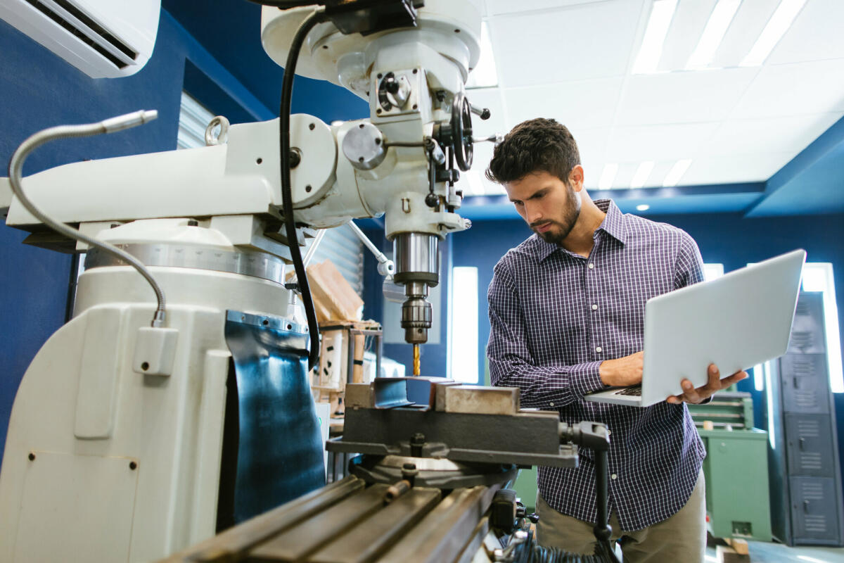 Ein junger lateinamerikanischer Ingenieur kalibriert eine große Bohrmaschine mit seinem Laptop in seiner Werkstatt. © aldomurillo/ Getty Images 