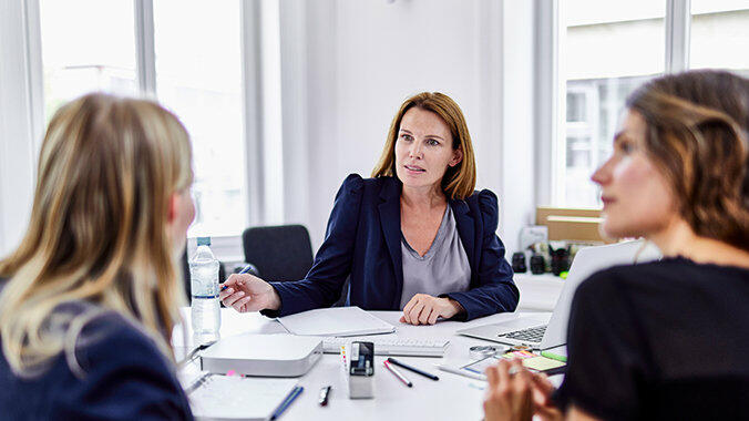 Drei Frauen sitzen an Schreibtisch und unterhalten sich © Oliver Rossi / Getty Images