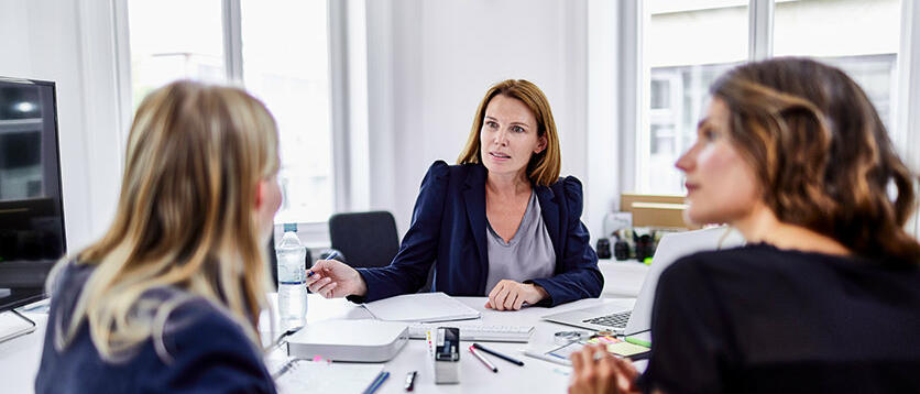 Drei Frauen sitzen an Schreibtisch und unterhalten sich © Oliver Rossi / Getty Images