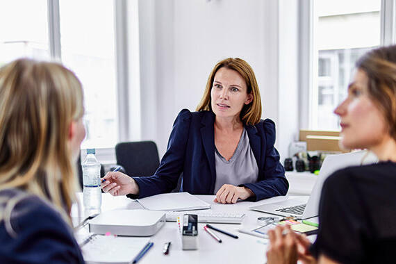 Drei Frauen sitzen an Schreibtisch und unterhalten sich © Oliver Rossi / Getty Images