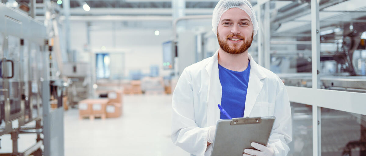 Gut aussehend lächelnd Nachwuchswissenschaftler mit Zwischenablage posiert in Fabrik  © Getty Images/ AleksandarGeorgiev