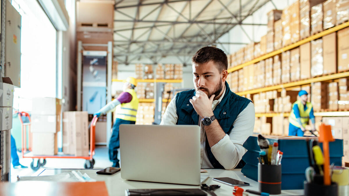 Der Supply Chain Manager sitzt am Laptop in einer Warenversandhalle © Marko Geber / Getty Images