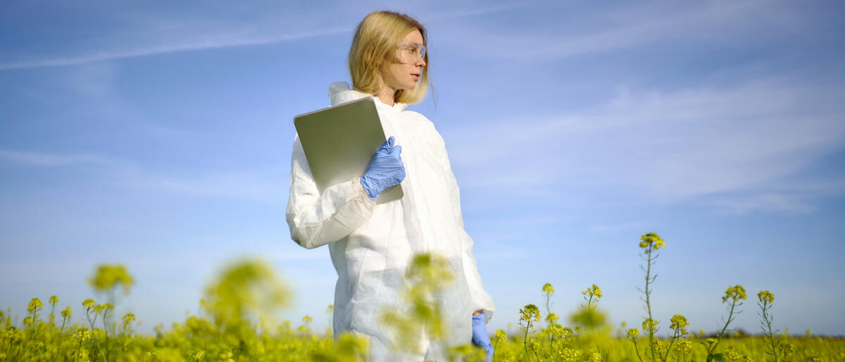 Eine junge Wissenschaftlerin steht im Schutzanzug mit einem Laptop in der Hand auf einem Feld. © Andriy Onufriyenko / Getty Images