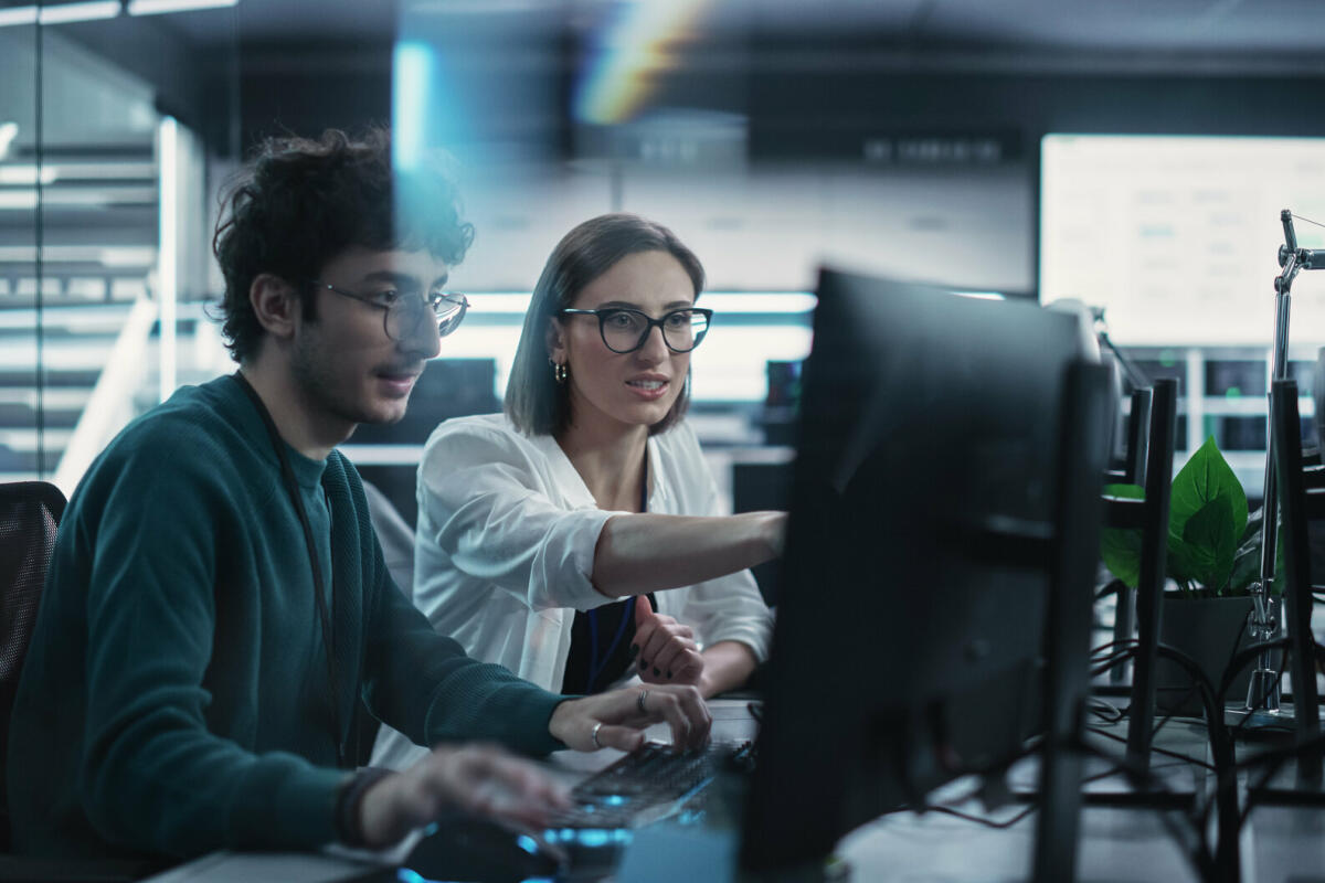 Zwei junge Kollegen, die am Computer arbeiten und sich am Arbeitsplatz unterhalten. Weibliche und männliche Softwareentwickler diskutieren eine Lösung. © gorodenkoff/ Getty Images