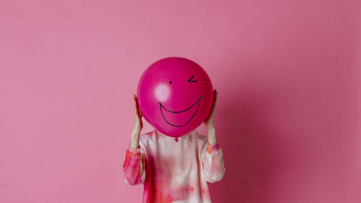 Auf rosa Hintergrund ist ein dreiviertellanges Studioporträt einer jungen Frau in Freizeitkleidung zu sehen. Sie hält einen rosa Ballon mit einem Lächeln. © SolStock / Getty Images