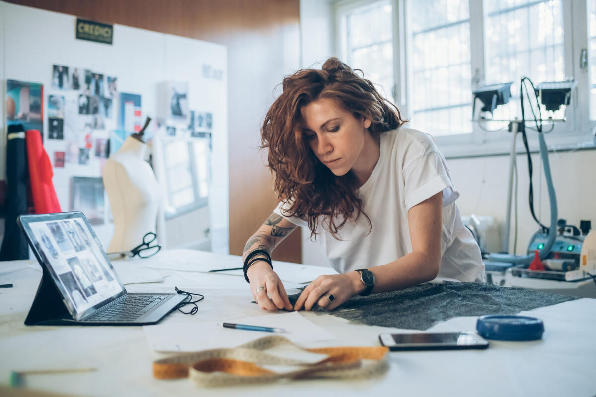 Eine junge tätowierte Textildesignerin beugt sich über ihren Arbeitstisch und arbeitet an einem Stück Stoff © Eugenio Marongiu / Getty Images