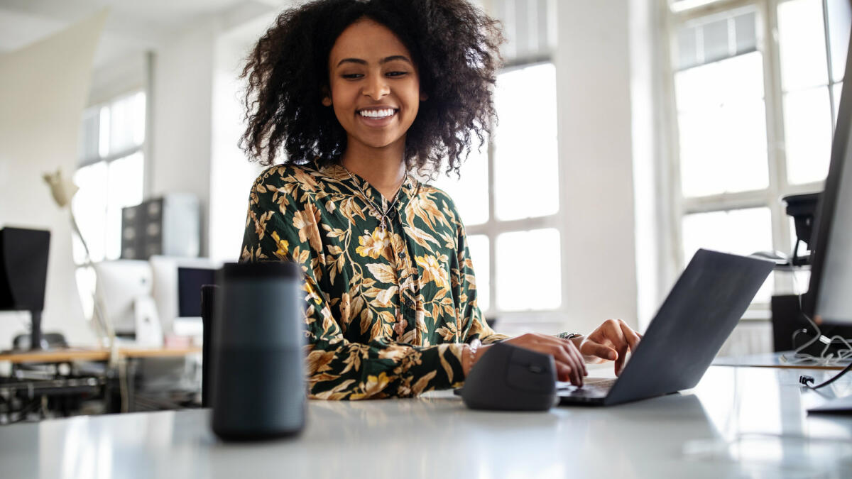 Lächelnde Frau sitzt vor Laptop und schaut Smart Speaker an © Luis Alvarez / Getty Images