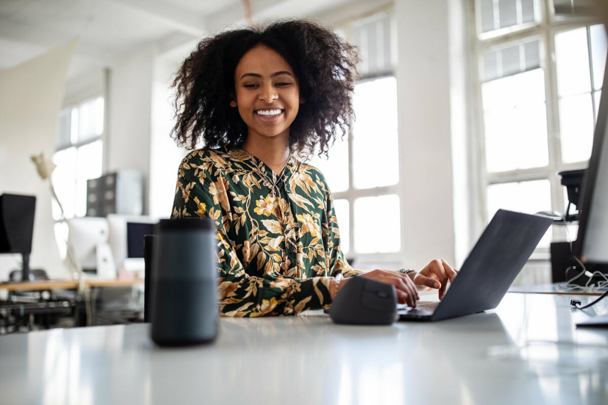 Lächelnde Frau sitzt vor Laptop und schaut Smart Speaker an © Luis Alvarez / Getty Images