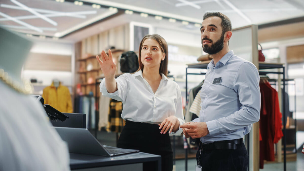 Bekleidungsgeschäft: Geschäftsfrau verwendet Laptop-Computer, spricht mit Visual Merchandising-Spezialisten und arbeiten zusammen. © Getty Images/ gorodenkoff