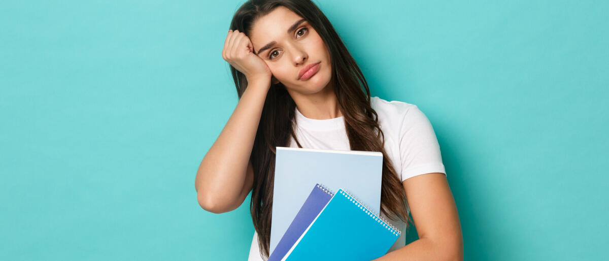 Porträt einer müden, hübschen Studentin, die Studienmaterial und Notizbücher in der Hand hält und erschöpft aussieht, stehend vor blauem Hintergrund © Mix and Match Studio / 500px / Getty Images