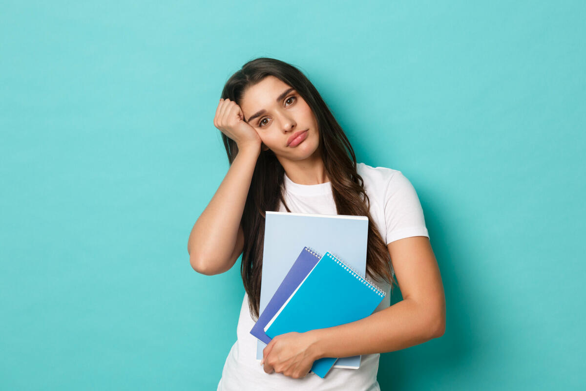 Porträt einer müden, hübschen Studentin, die Studienmaterial und Notizbücher in der Hand hält und erschöpft aussieht, stehend vor blauem Hintergrund © Mix and Match Studio / 500px / Getty Images