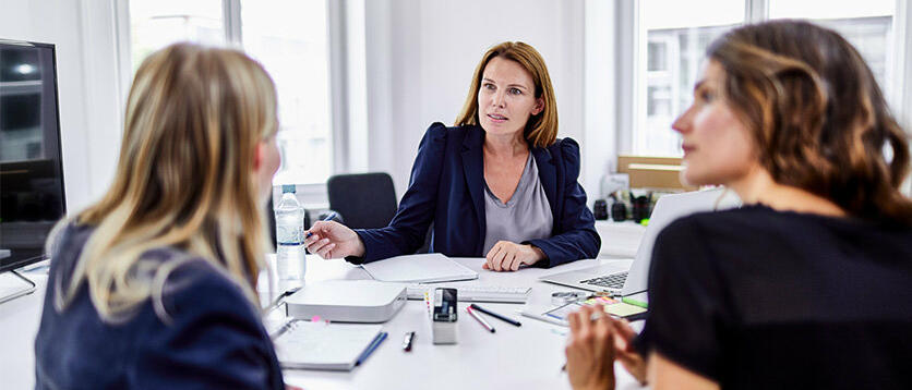 Drei Frauen sitzen an einem Schreibtisch und sehen sich an. © Oliver Rossi / Getty Images