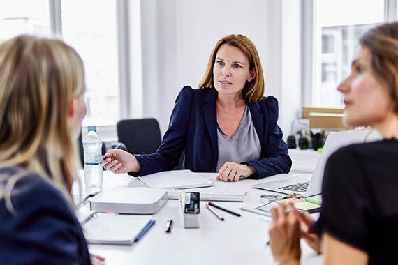 Drei Frauen sitzen an einem Schreibtisch und sehen sich an. © Oliver Rossi / Getty Images