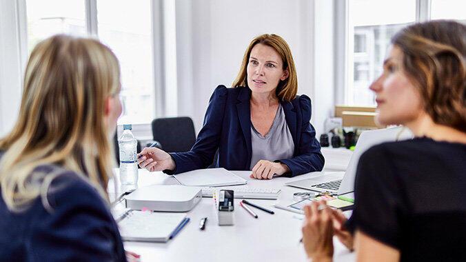 Drei Frauen sitzen an einem Schreibtisch und sehen sich an. © Oliver Rossi / Getty Images