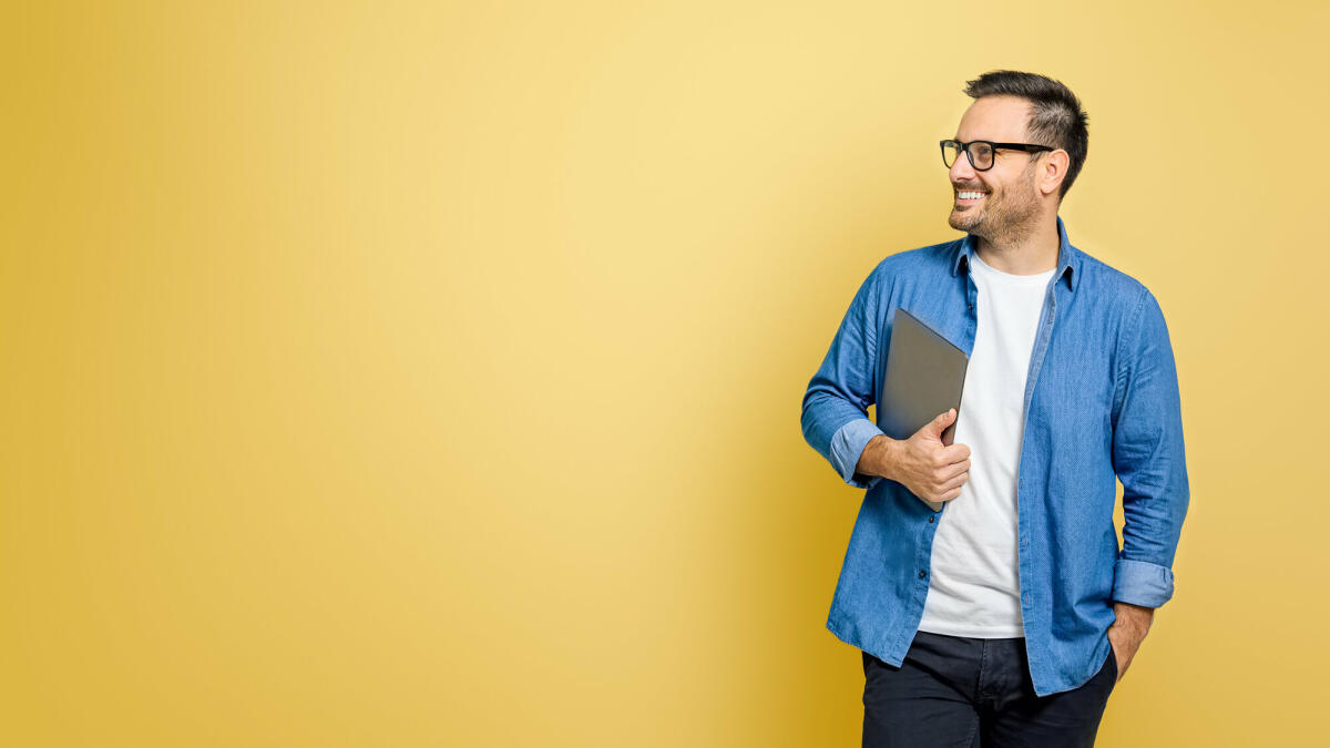 Glücklicher männlicher Freiberufler mit Hand in der Tasche, der digitales Tablet hält und über gelben Hintergrund wegschaut © Moon Safari / Getty Images