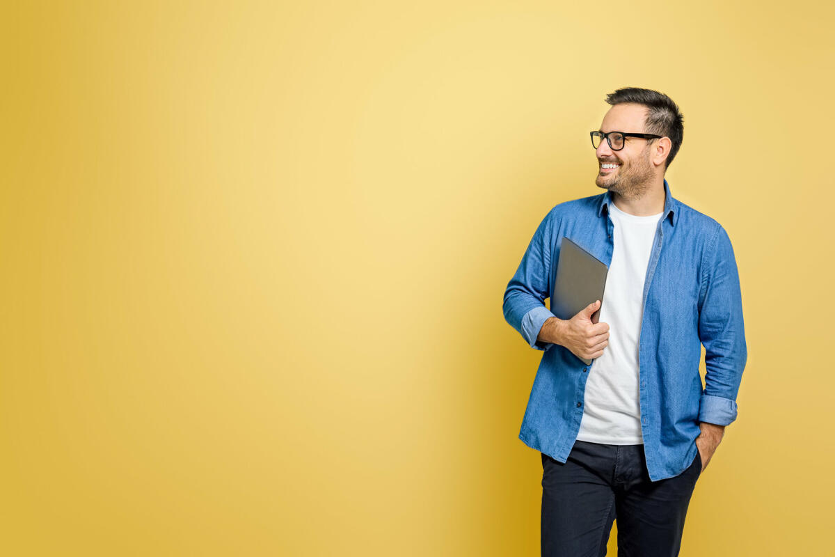 Glücklicher männlicher Freiberufler mit Hand in der Tasche, der digitales Tablet hält und über gelben Hintergrund wegschaut © Moon Safari / Getty Images