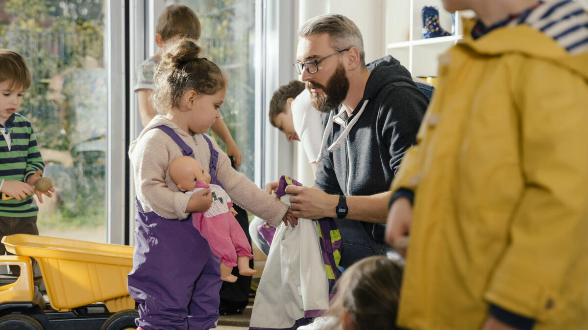 Erzieher hilft Mädchen beim Anziehen der Regenkleidung im Kindergarten. © Westend61/ Getty Images