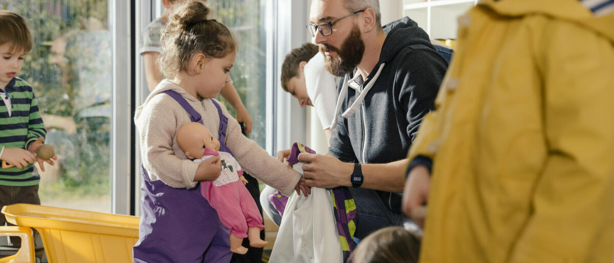 Erzieher hilft Mädchen beim Anziehen der Regenkleidung im Kindergarten. © Westend61/ Getty Images