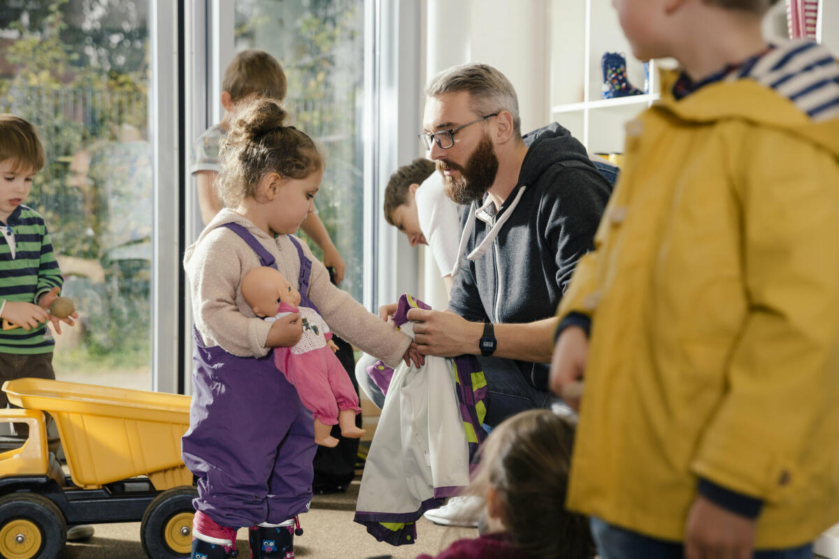 Erzieher hilft Mädchen beim Anziehen der Regenkleidung im Kindergarten. © Westend61/ Getty Images