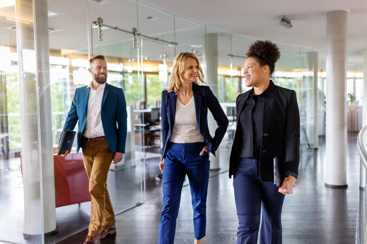 Zwei Frauen und ein Mann in Business-Kleidung gehen durchs Büro. © Luis Alvarez / Getty Images