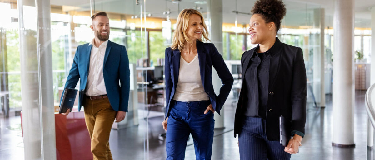 Zwei Frauen und ein Mann in Business-Kleidung gehen durchs Büro. © Luis Alvarez / Getty Images