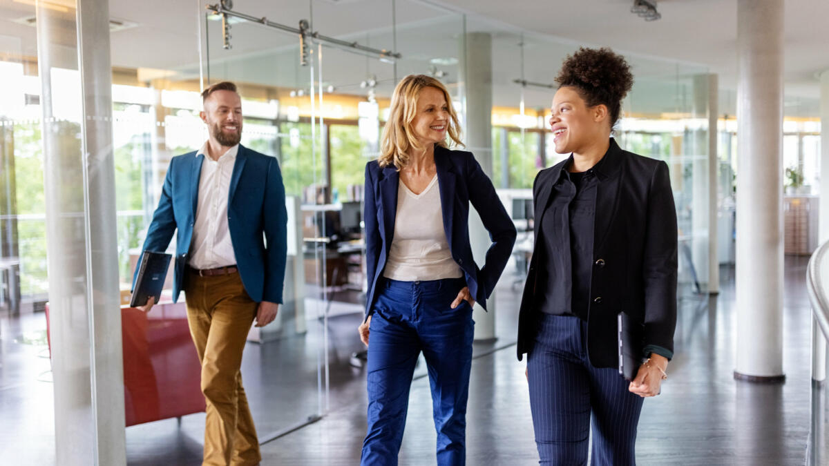 Zwei Frauen und ein Mann in Business-Kleidung gehen durchs Büro. © Luis Alvarez / Getty Images