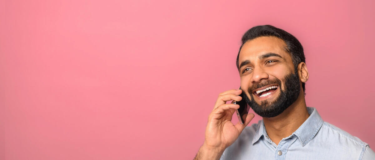 Fröhlicher optimistischer junger indischer bärtiger Mann, der telefoniert, um Pläne zu besprechen, und mit verträumtem Gesichtsausdruck wegschaut. © Vadym Pastukh / Getty Images