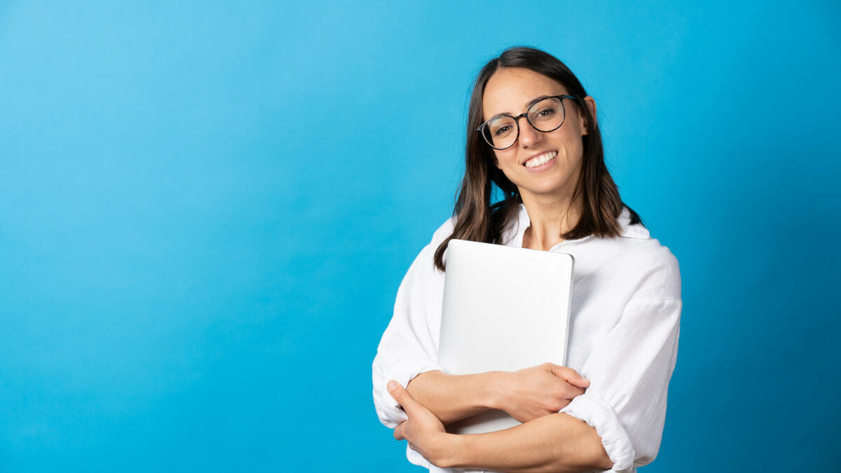 Hübsche hispanische Frau, die einen Laptop hält und in die Kamera schaut © Egoitz Bengoetxea Iguaran / Getty Images