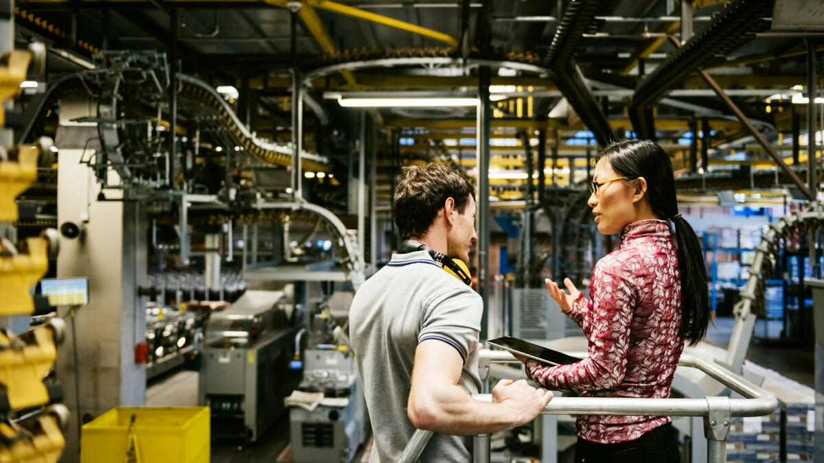 Mechaniker und Manager sprechen in der Nähe einer Maschine in einer großen Druckerei über den neuesten Zeitungsdruck © TommL/ Getty Images