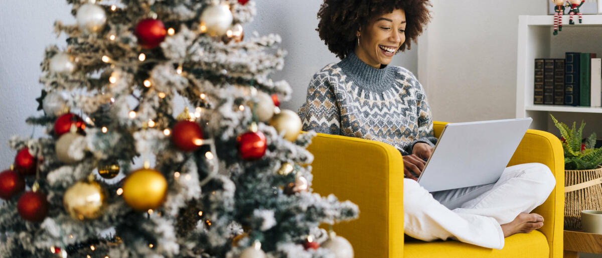 Frau mit Laptop sitzt auf Sessel neben Weihnachtsbaum © Westend61 / Getty Images