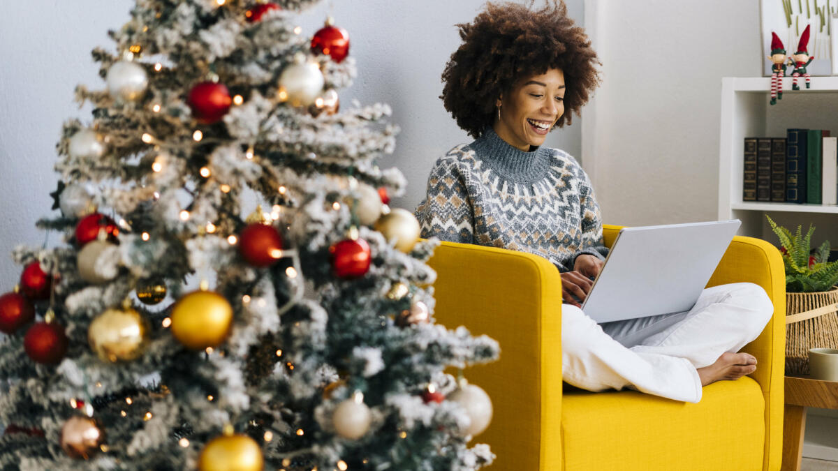 Frau mit Laptop sitzt auf Sessel neben Weihnachtsbaum © Westend61 / Getty Images