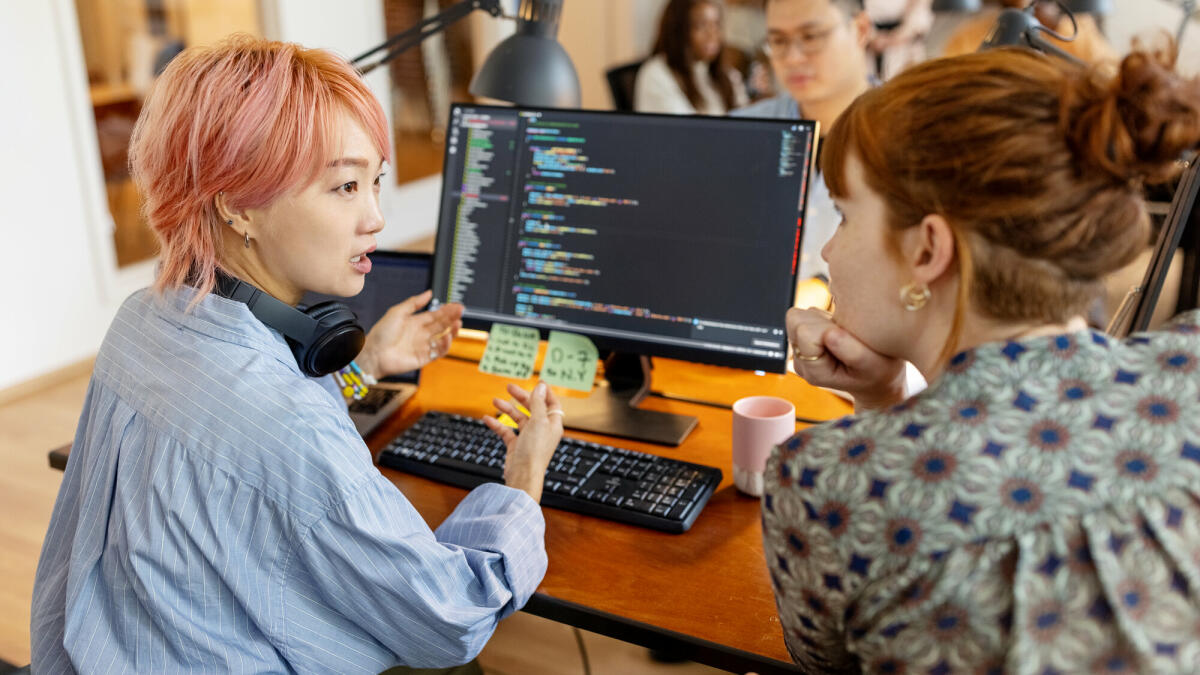 Weibliche Softwareentwicklerinnen sitzen am Arbeitsplatz am Computer und diskutieren kreativ über das neue Programmierprogramm im Büro. © Luis Alvarez/ Getty Images