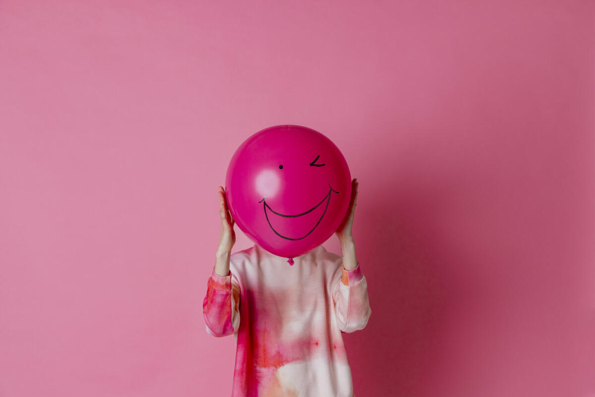 Frau hält einen rosa Ballon mit einem Lächeln auf dem Ballon vor ihrem Gesicht © SolStock / Getty Images