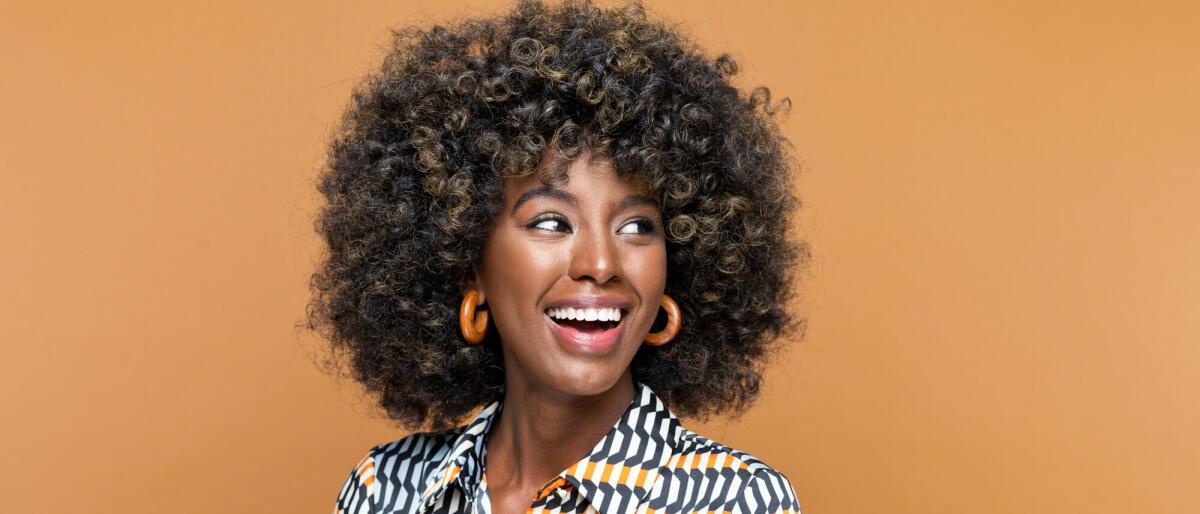 Erstaunte junge Frau mit Afrofrisur, die ein bedrucktes Kleid und hölzerne Ohrringe trägt, wegschaut und lacht. Studioaufnahme auf braunem Hintergrund © izusek / Getty Images