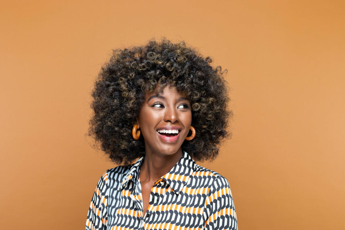 Erstaunte junge Frau mit Afrofrisur, die ein bedrucktes Kleid und hölzerne Ohrringe trägt, wegschaut und lacht. Studioaufnahme auf braunem Hintergrund © izusek / Getty Images