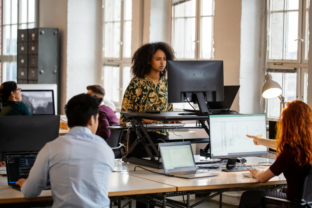 Junge afrikanische Frau arbeitet an einem Stehpult im Büro. Weibliche Mitarbeiterin arbeitet am Computer an einem ergonomischen Stehpult. © Luis Alvarez/ Getty Images