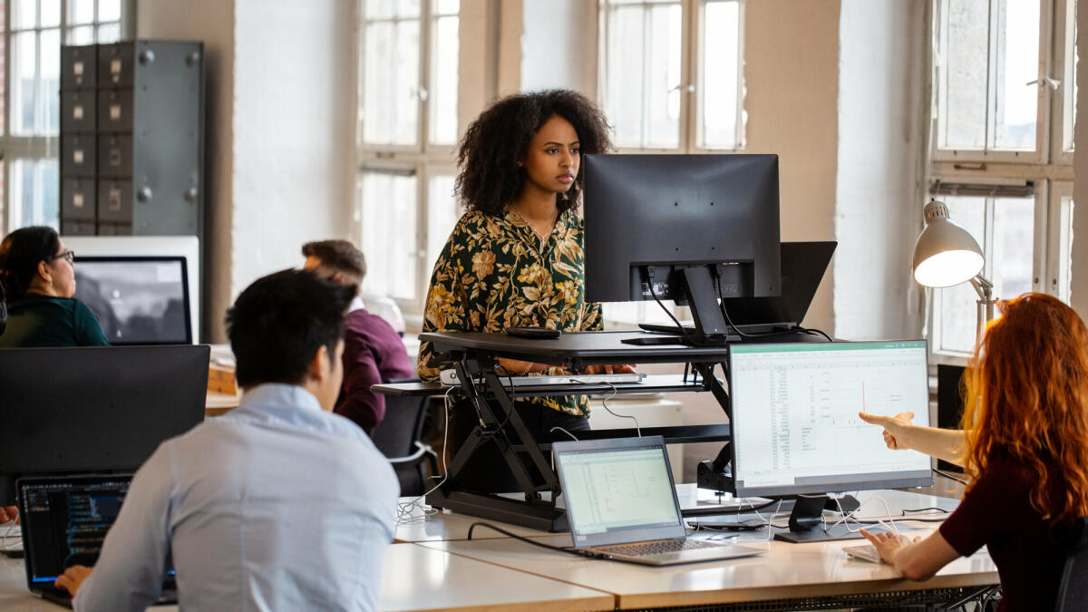 Junge afrikanische Frau arbeitet an einem Stehpult im Büro. Weibliche Mitarbeiterin arbeitet am Computer an einem ergonomischen Stehpult. © Luis Alvarez/ Getty Images