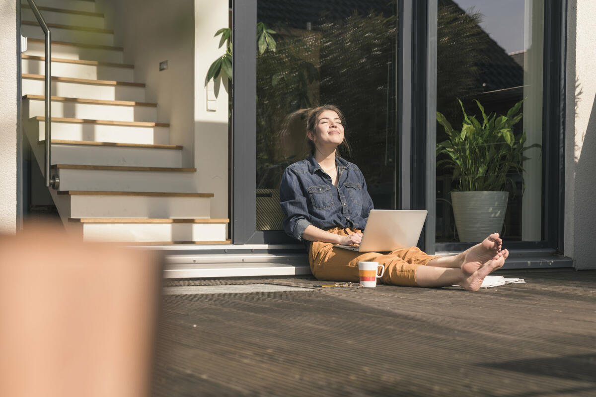 Junge Frau mit Laptop auf dem Schoß sitzt mit geschlossenen Augen in der Sonne  © Westend61 / Getty Images