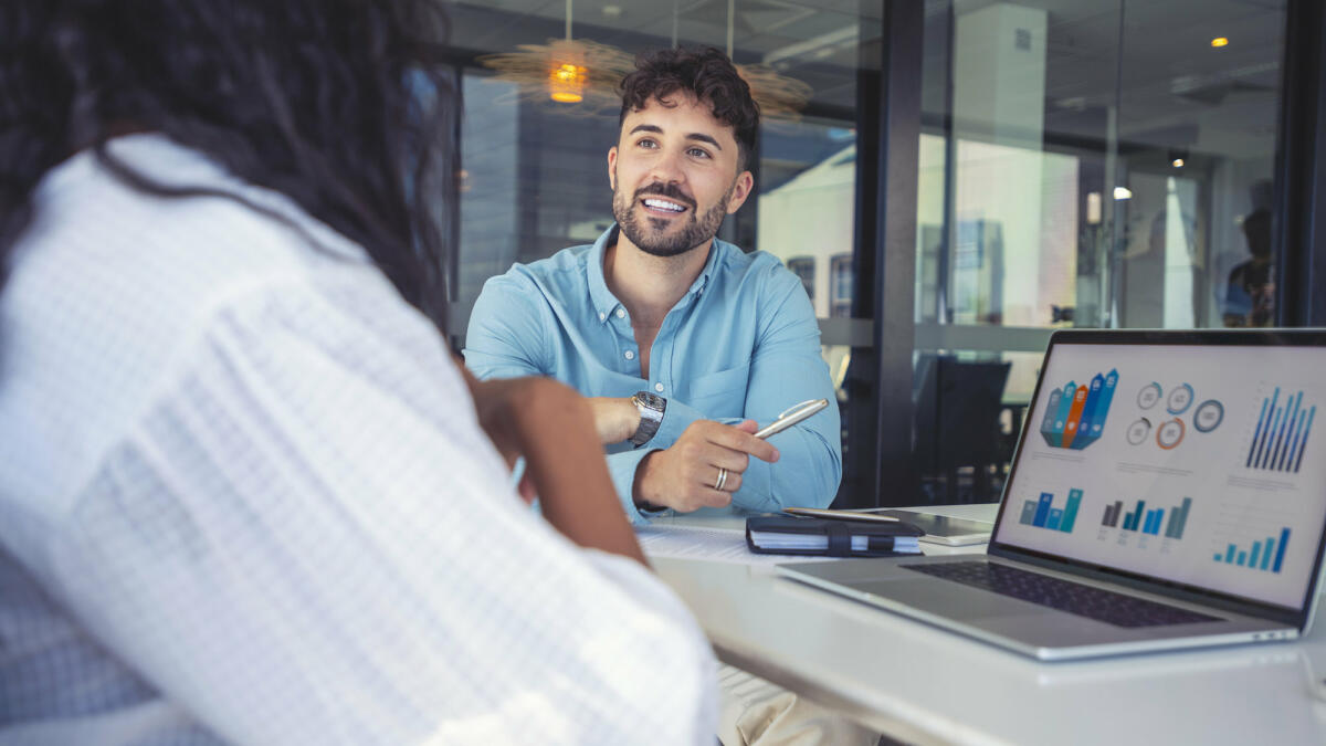 Ein junger Technologieberater sitzt zusammen mit der einer Klientin im Meetingroom und präsentiert Grafiken © courtneyk / Getty Images